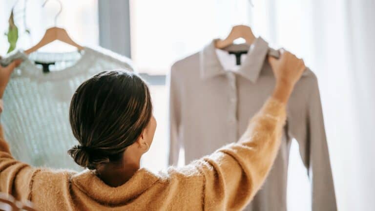 A person with dark hair in a yellow or mustard-colored sweater reaching up to hang or arrange a white shirt on a clothing rack or closet rod