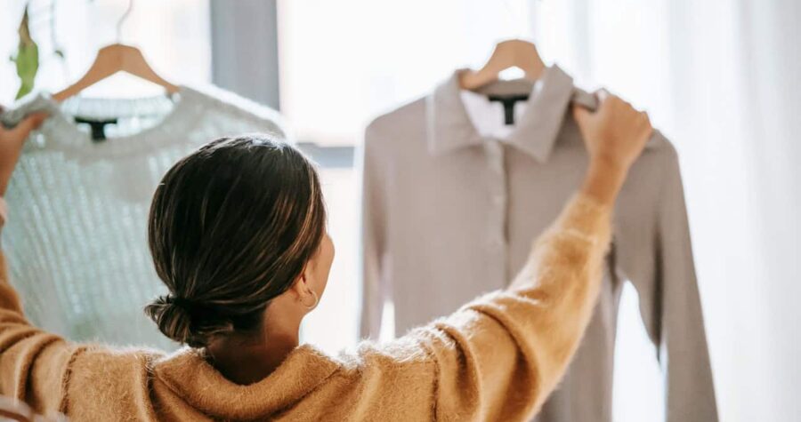 A person with dark hair in a yellow or mustard-colored sweater reaching up to hang or arrange a white shirt on a clothing rack or closet rod