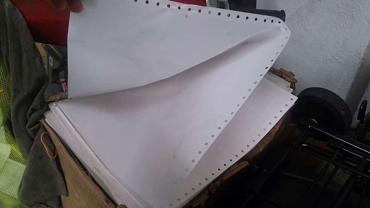 ontinuous-feed dot matrix printer paper with perforated edges and sprocket holes, stacked in a cardboard box in a storage or workshop setting
