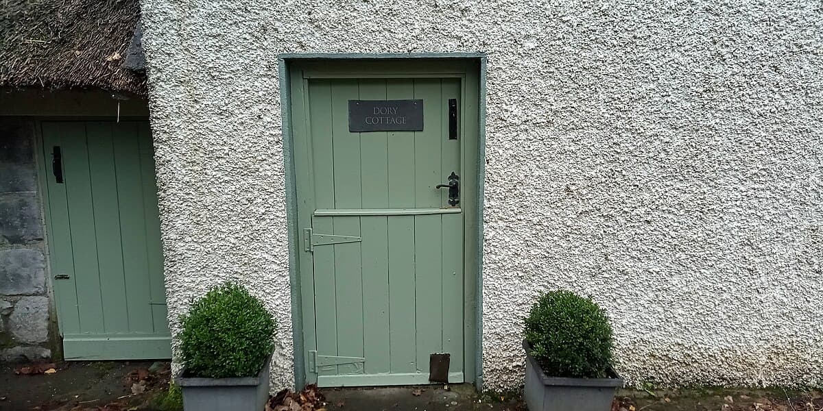 Green Dutch door, labeled “Dory Cottage,” white textured wall, potted shrubs on both sides