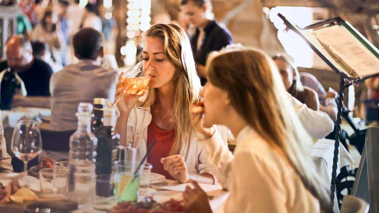 Two women dining at a busy restaurant, one drinking from a wine glass, the other eating pizza, surrounded by plates, glasses, and bottles