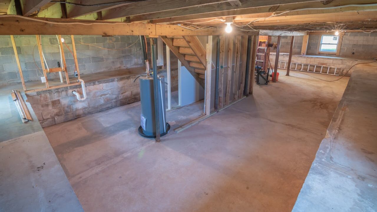 Unfinished basement with concrete floor, exposed wooden beams, cinder block walls, water heater, wooden stairs, and rough framing for future rooms