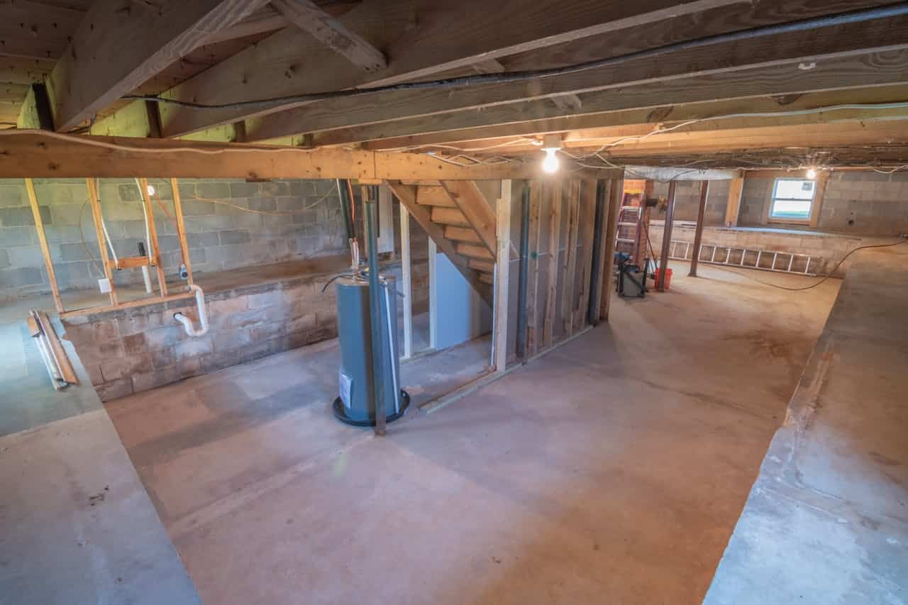 Unfinished basement with concrete floor, exposed wooden beams, cinder block walls, water heater, wooden stairs, and rough framing for future rooms
