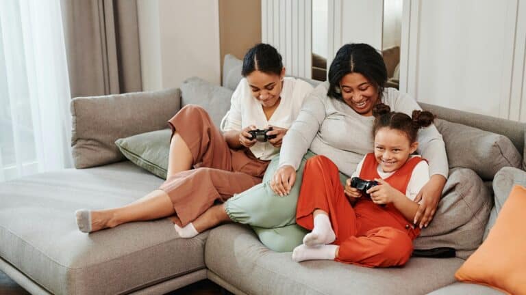 Two people sitting on a beige sofa using mobile devices or tablets, with one person wearing white and the other in orange or red clothing, in a modern living room setting