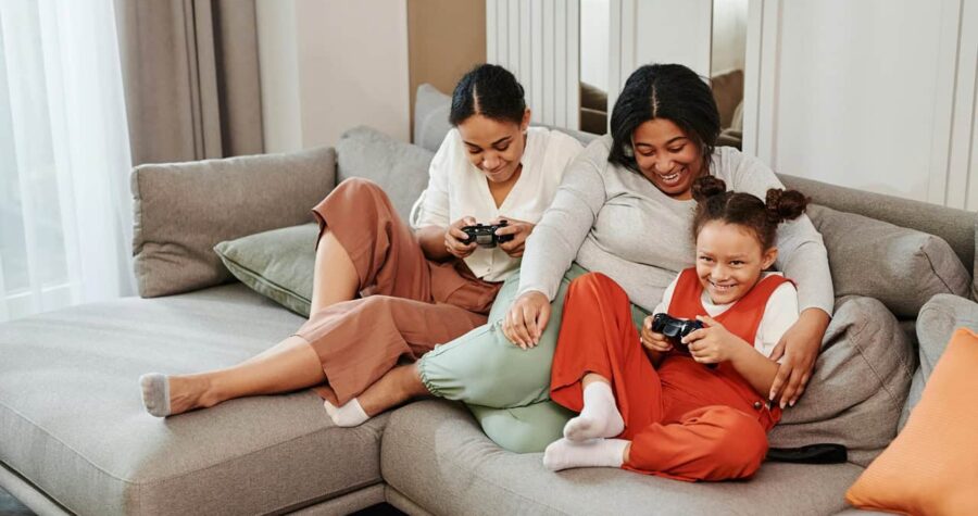 Two people sitting on a beige sofa using mobile devices or tablets, with one person wearing white and the other in orange or red clothing, in a modern living room setting