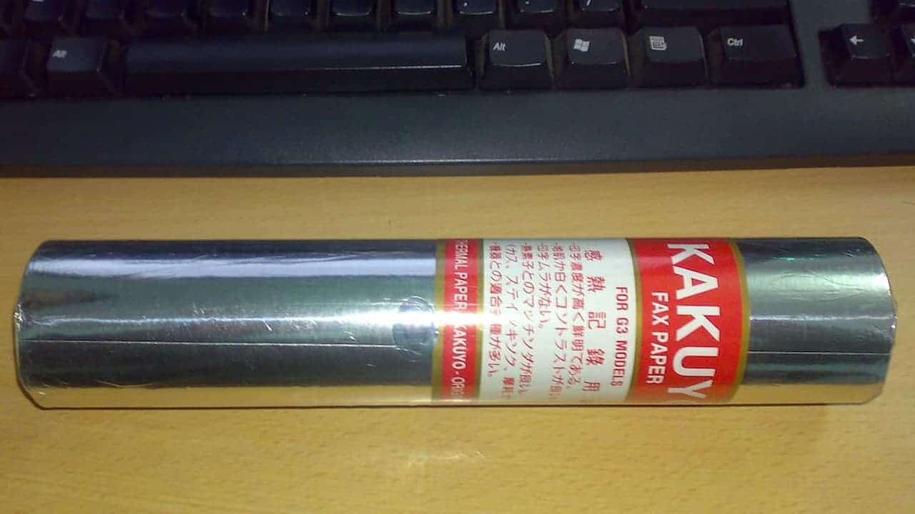 Roll of KAKUYO fax paper wrapped in shiny metallic packaging with red and white labeling, placed on a wooden desk in front of a black computer keyboard