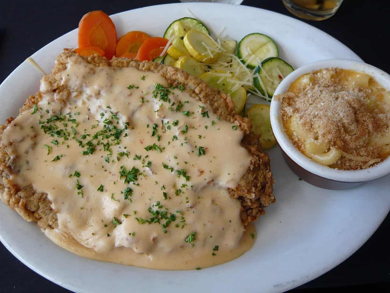 Plate of chicken fried steak topped with creamy gravy and chopped parsley, served with sautéed zucchini and carrots, and a side of baked macaroni and cheese in a small bowl, arranged on a white oval plate for a hearty southern-style meal