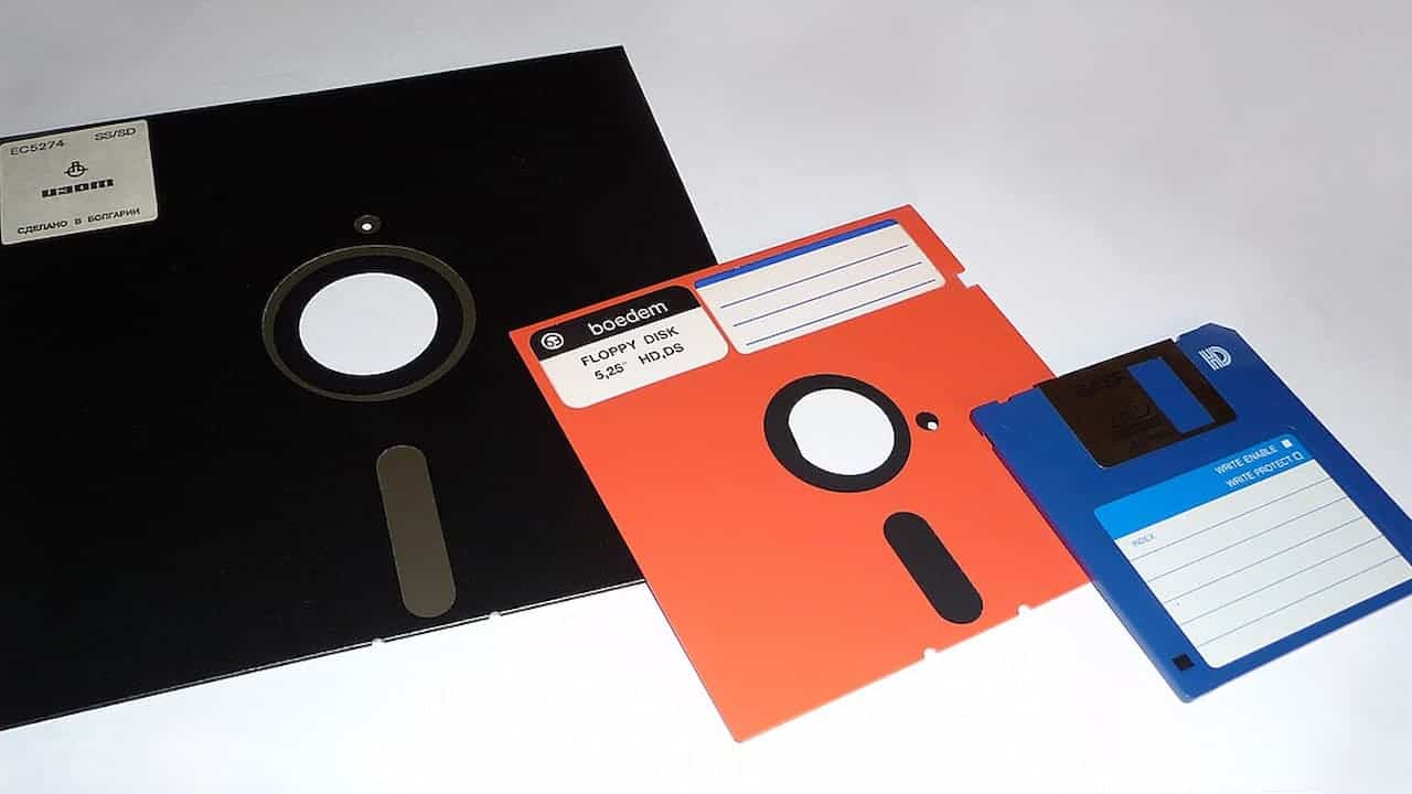Three floppy disks of different sizes and colors displayed side by side—a large black 8-inch disk, a red 5.25-inch disk, and a blue 3.5-inch disk with a metal shutter