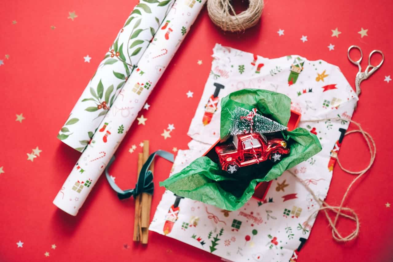 Christmas-themed gift wrapping scene with red toy truck carrying a tree in green tissue paper, rolls of wrapping paper, ribbon, twine