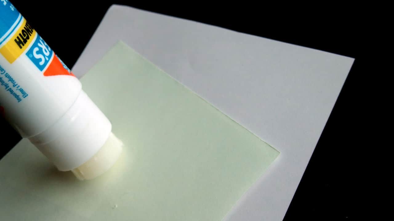 Half-used glue stick applying adhesive to a light green sheet on top of a white paper, set against a black background