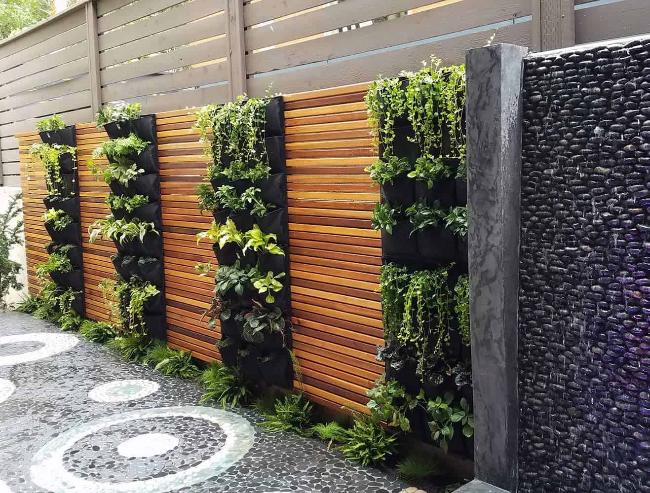 Modern garden wall featuring vertical plant pockets on wooden slat fence, black stone water feature, decorative pebble pathway with circular pattern