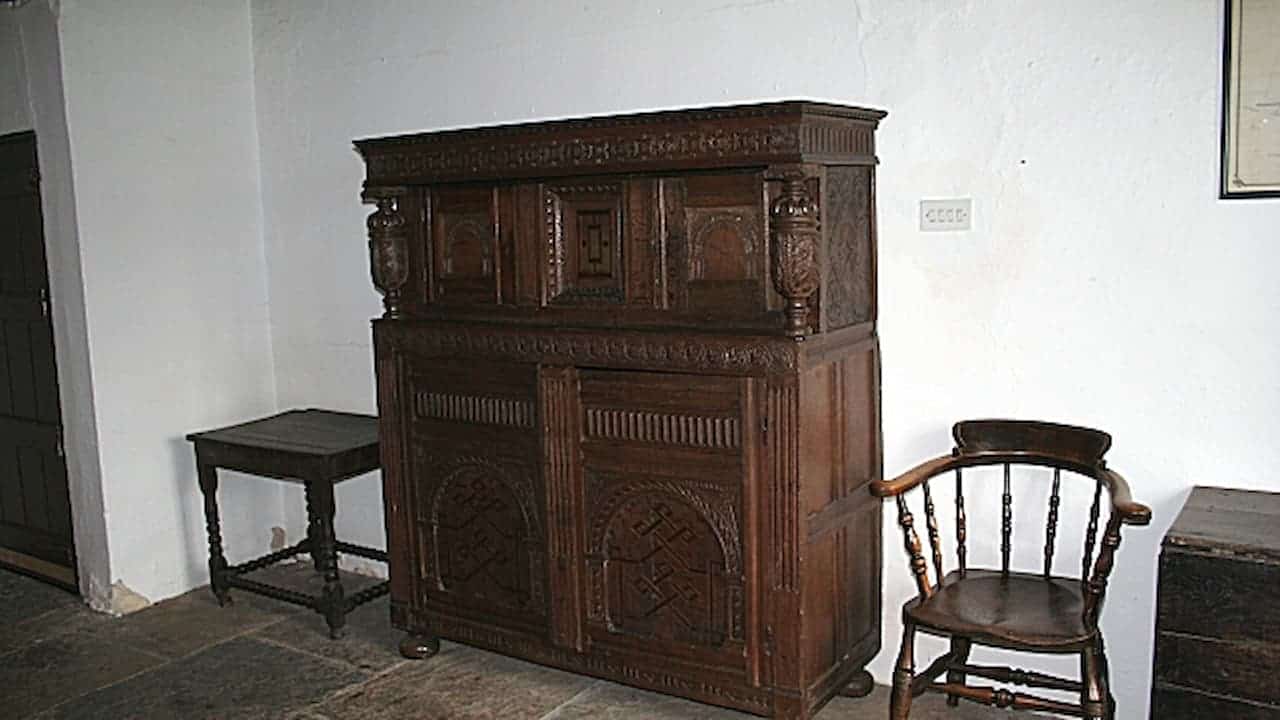 Ornate carved oak cabinet with decorative panels displayed in museum room alongside wooden chair and side table