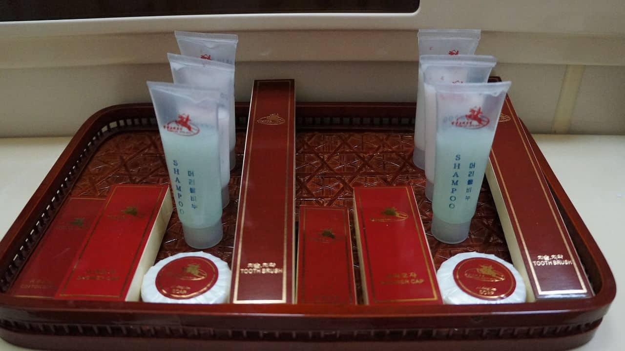 Neatly arranged hotel toiletries on a decorative tray, including shampoo tubes, toothbrush kits, soaps, shower caps, and vanity kits in red and gold packaging