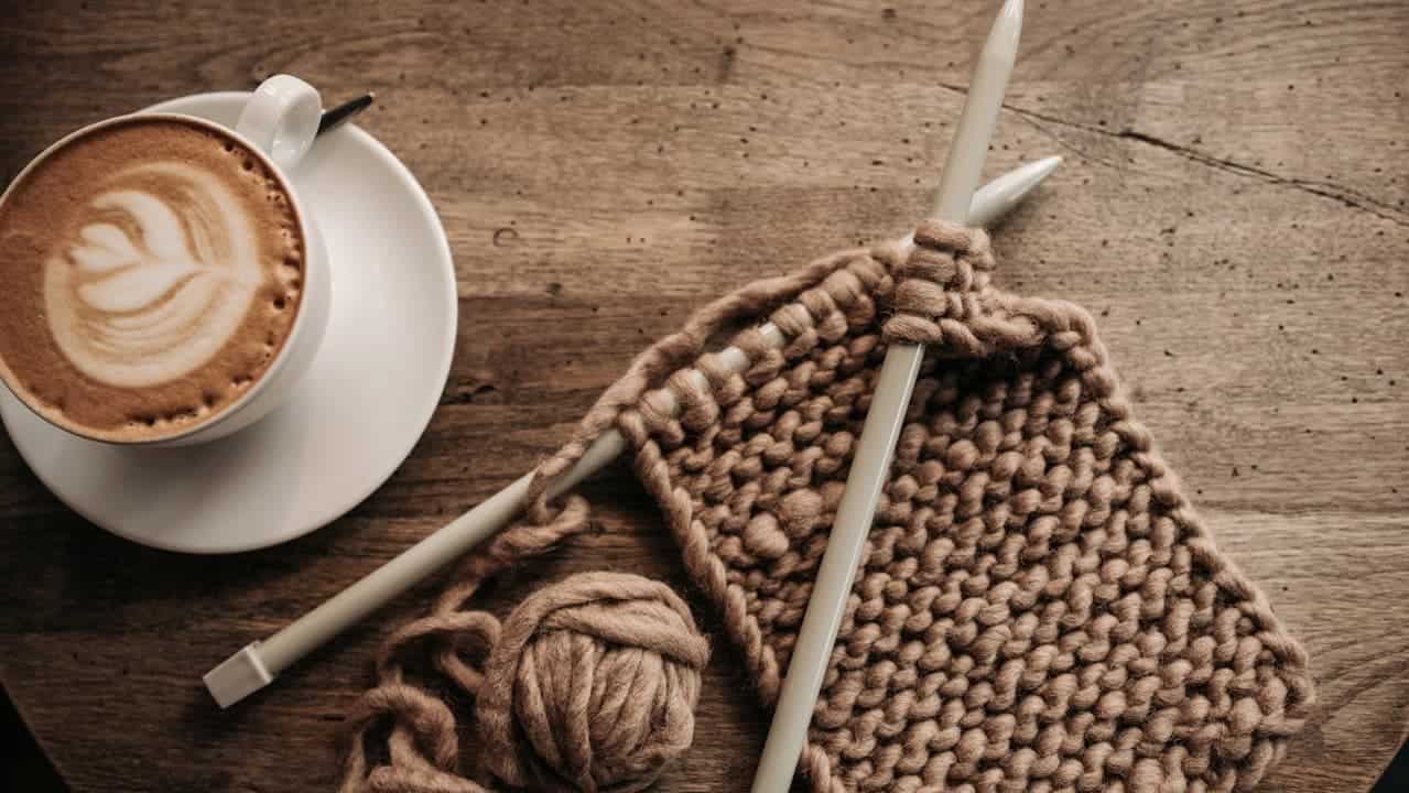 Latte with leaf pattern, brown knitting project on needles, yarn ball on rustic wooden table