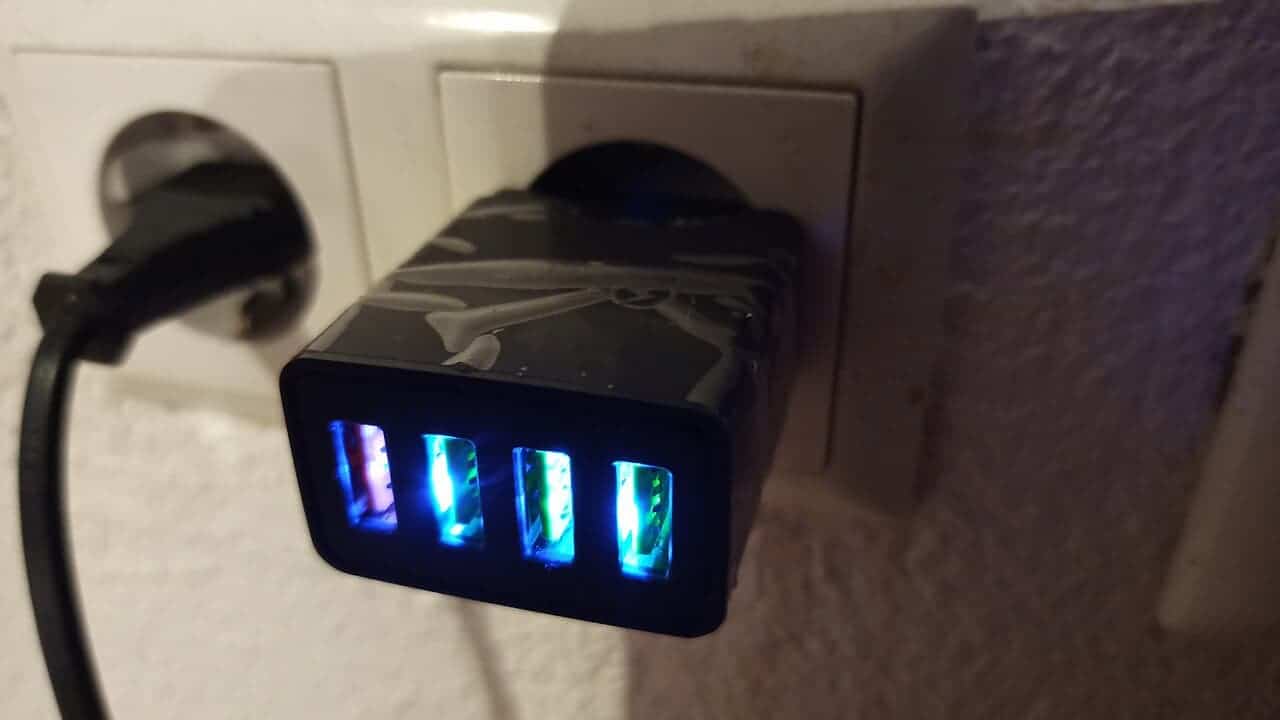 A black USB wall charging brick with four illuminated USB ports plugged into a wall socket, showing a blue glow