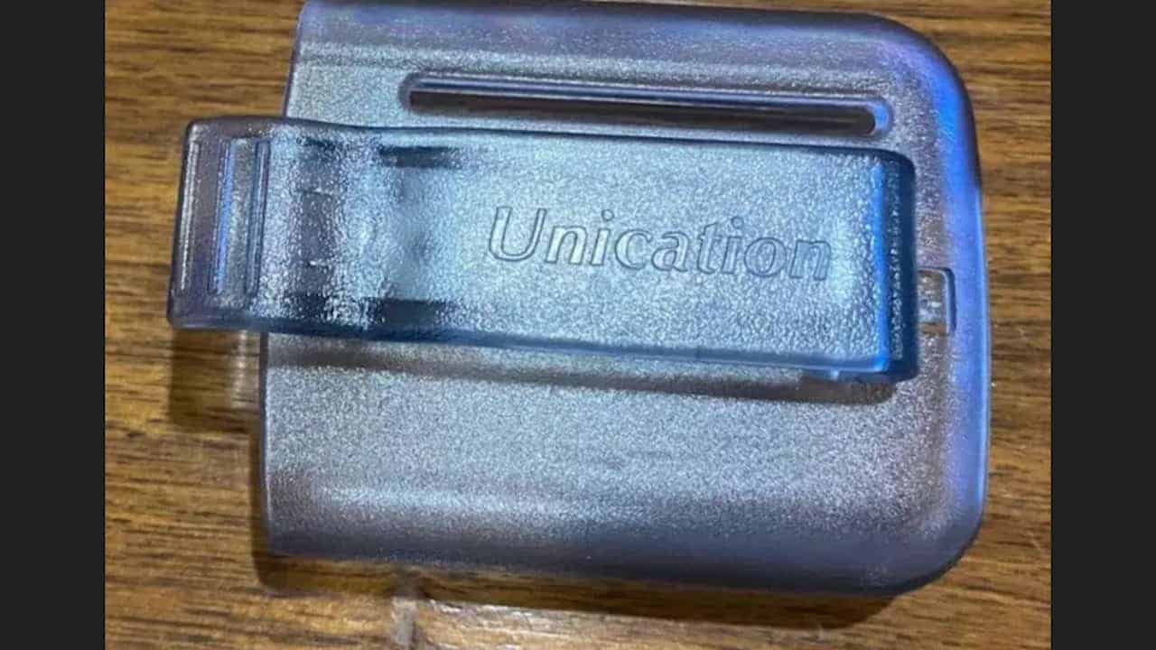  A translucent plastic pager belt clip labeled “Unication” resting on a wooden surface, designed to hold a pager securely on a belt
