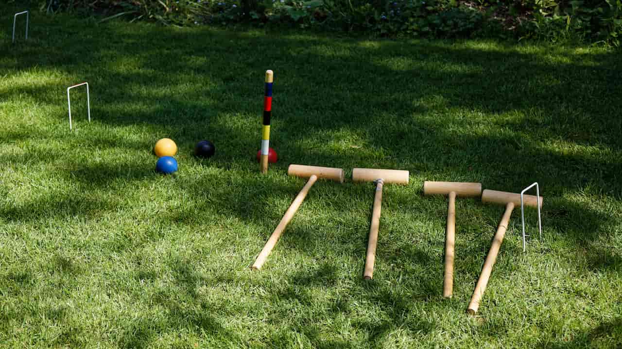 A croquet set arranged on a grassy lawn with four wooden mallets, colorful balls, metal hoops, and a striped center peg