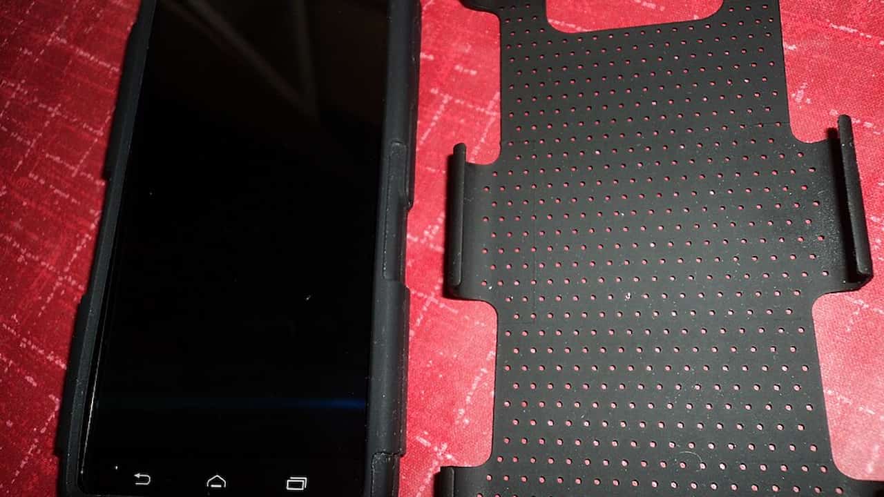 A black smartphone with a rugged protective case next to a separate perforated plastic back cover, both placed on a red patterned surface