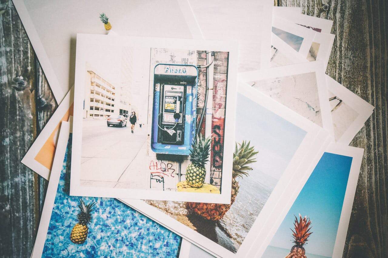Scattered stack of vibrant postcards on a wooden surface, top card features a street phone booth with pineapples, others show beach scenes and pineapples in creative settings