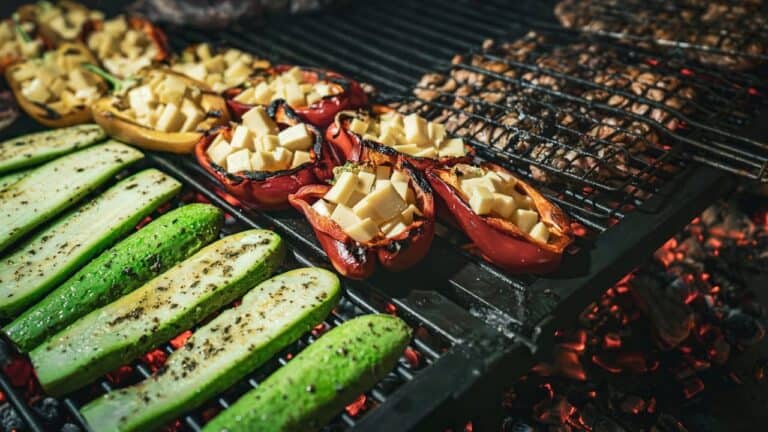 Grilled vegetables on barbecue grill, including zucchini slices, stuffed red bell peppers with cheese, over glowing coals
