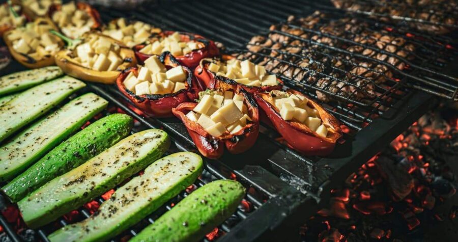 Grilled vegetables on barbecue grill, including zucchini slices, stuffed red bell peppers with cheese, over glowing coals