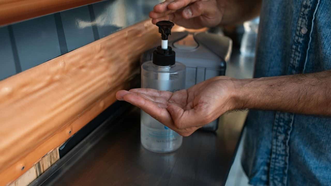 Person dispensing rubbing alcohol from clear pump bottle into palm, wooden counter edge visible, wearing denim clothing, in indoor setting