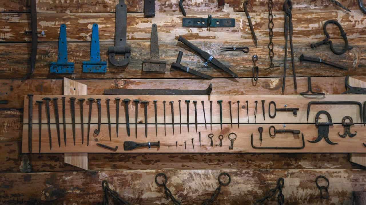 Vintage blacksmith tools displayed on weathered wooden wall, including hammers, tongs, chisels, measuring tools, chains, and metal implements