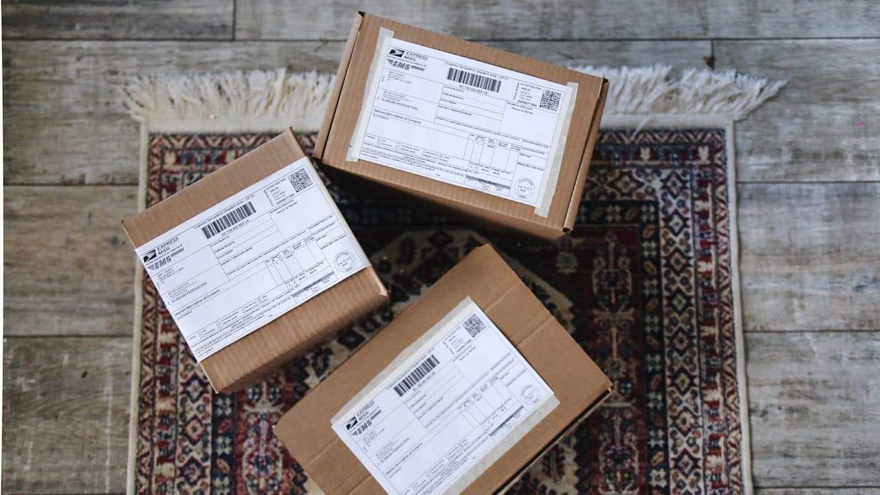 Three cardboard packages with USPS shipping labels, placed on a patterned rug, wooden floor background