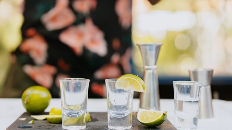 Three clear shot glasses with lime wedges on a cutting board, surrounded by fresh lime slices and metal jiggers, person in floral shirt in background