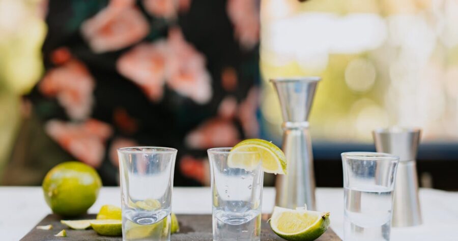 Three clear shot glasses with lime wedges on a cutting board, surrounded by fresh lime slices and metal jiggers, person in floral shirt in background