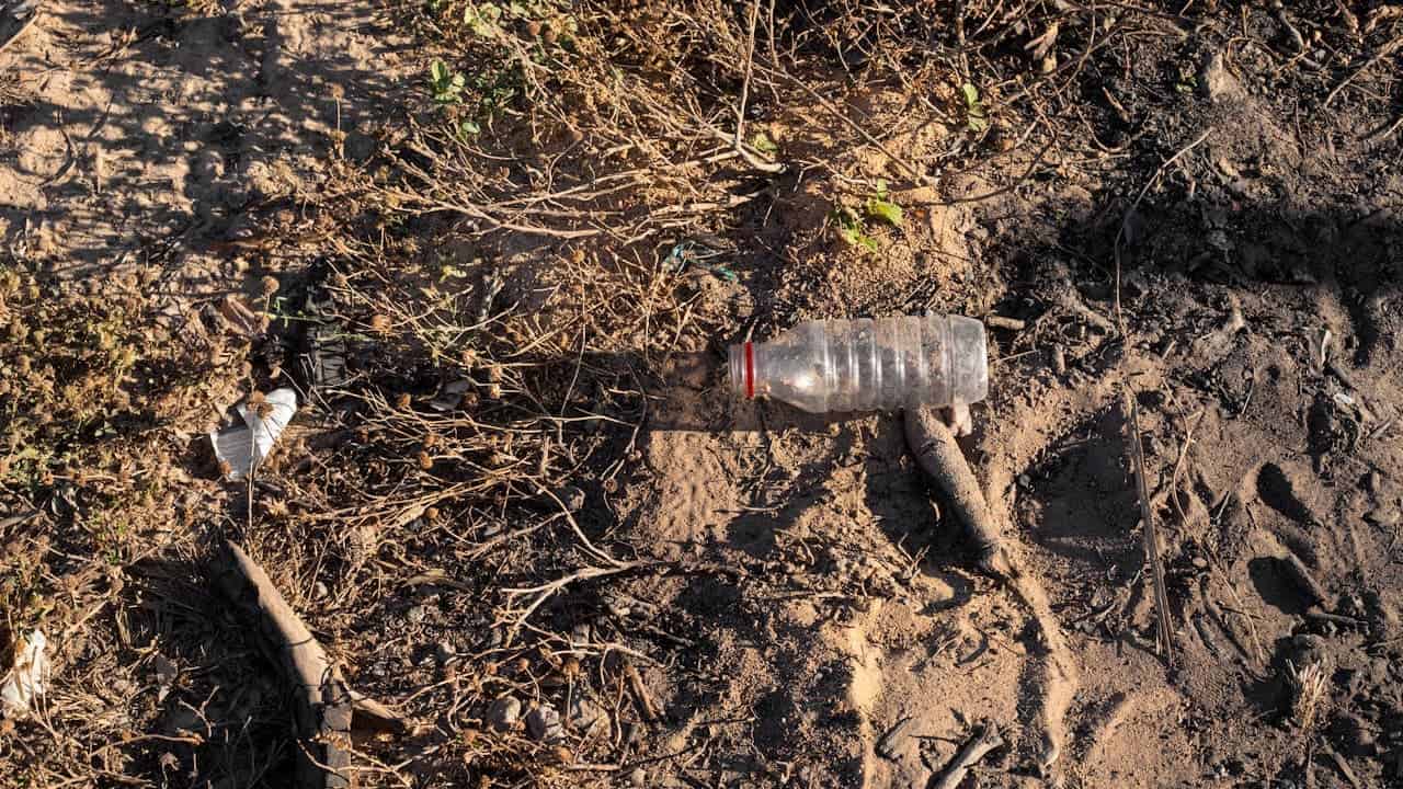 Discarded plastic water bottle lying on dry, sandy soil, surrounded by dead vegetation, branches, and other litter, showing environmental pollution in arid landscape