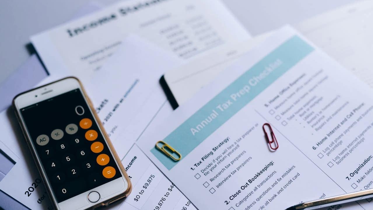 Calculator app on smartphone displaying numbers, scattered financial documents and tax forms on white surface, red paperclip attached to papers