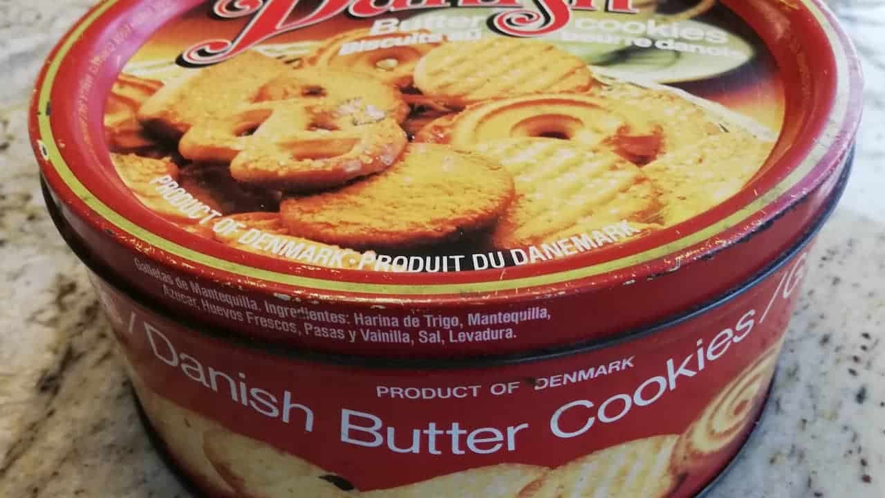Round red tin of Danish butter cookies with images of assorted cookies on the lid, labeled “Product of Denmark” in English and French, placed on a speckled kitchen countertop