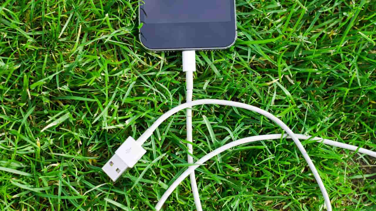 A smartphone connected to white USB charging cables lying on green grass, with the USB plug visible and the phone screen turned off