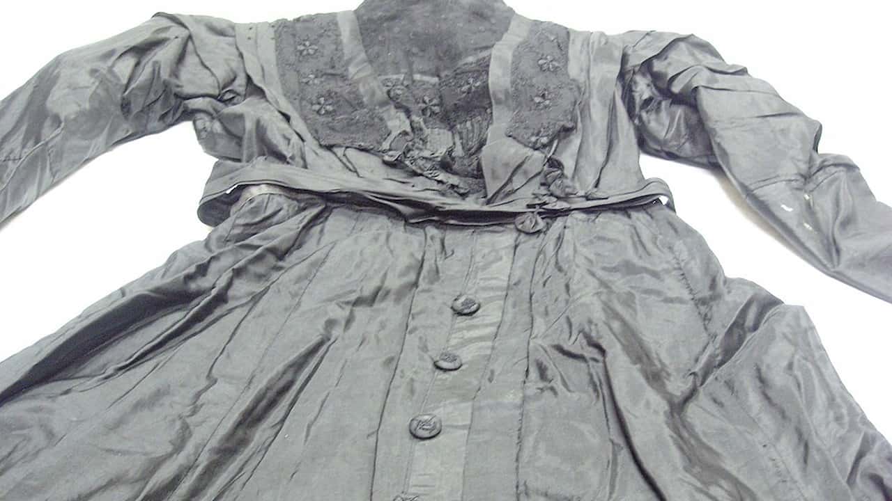 Worn-out vintage black dress with long sleeves and decorative lace on the chest, displayed flat on a white background, buttons running down the front