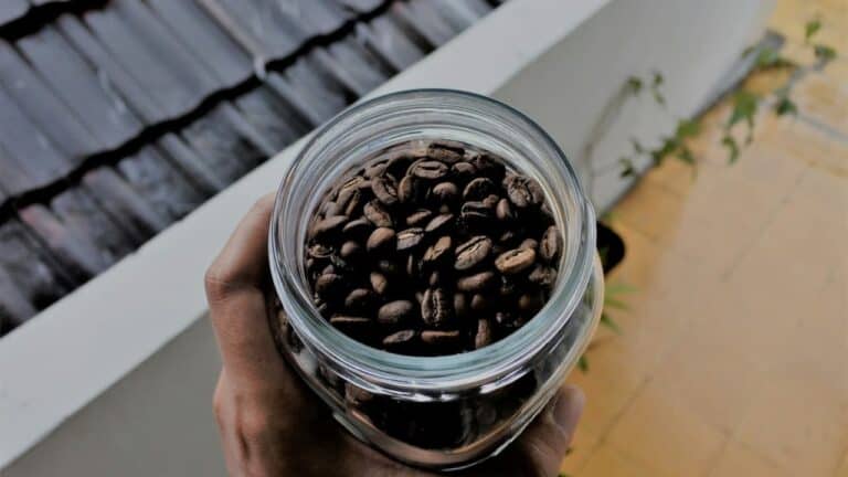 Hand holding a glass jar filled with roasted coffee beans, viewed from above, standing near a tiled floor and outdoor wall, natural lighting highlighting the rich brown tones of the beans