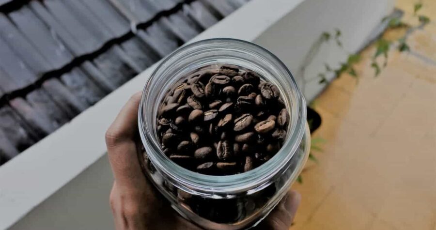 Hand holding a glass jar filled with roasted coffee beans, viewed from above, standing near a tiled floor and outdoor wall, natural lighting highlighting the rich brown tones of the beans