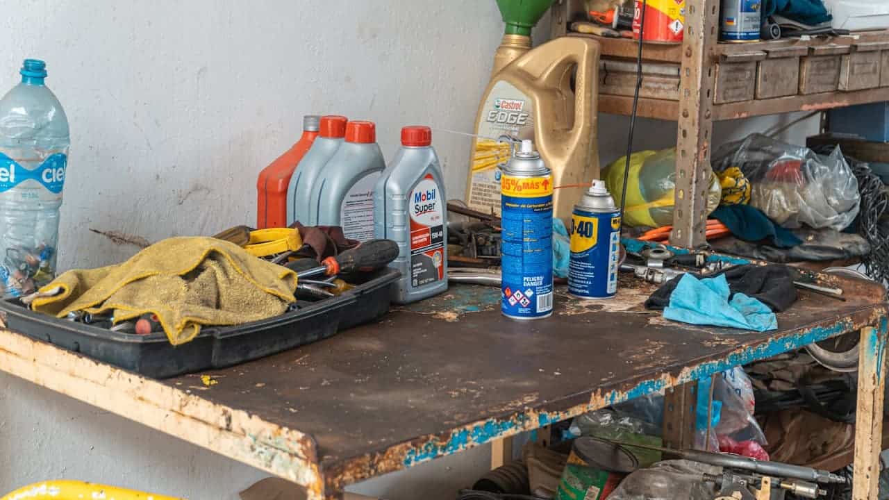 Cluttered garage workbench with motor oil bottles, Castrol Edge container, WD-40 spray, dirty rags, tools, shelving with miscellaneous items