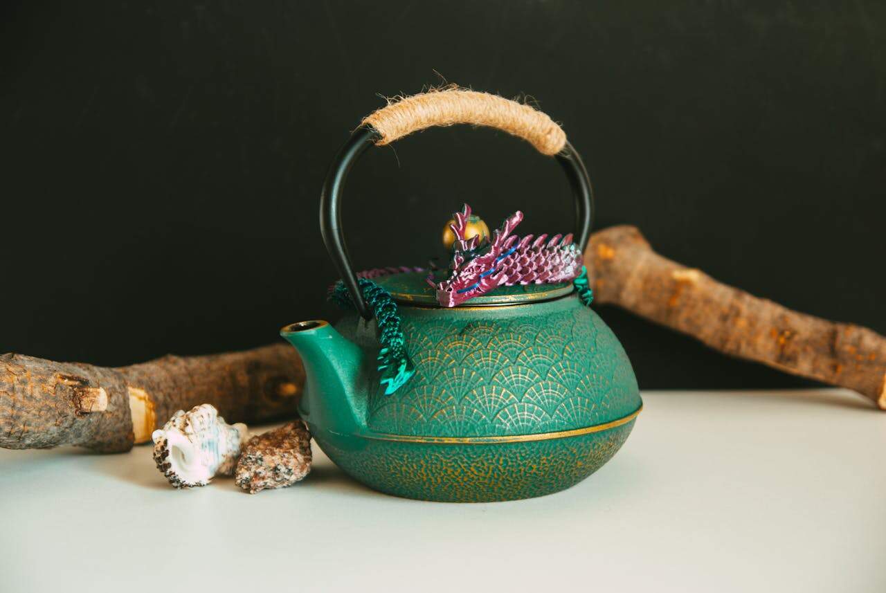 Ornate green teapot with gold pattern, rope handle, colorful dragon figurine on lid, displayed with driftwood and seashells on white surface