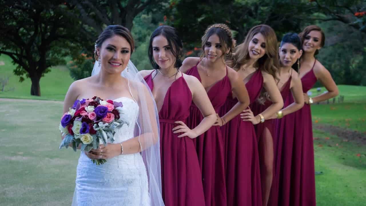 Bride in white dress with colorful bouquet poses with five bridesmaids in burgundy dresses, outdoor park setting