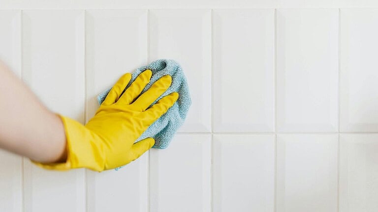 Person wearing a yellow rubber glove wiping white tiled wall with a blue microfiber cloth