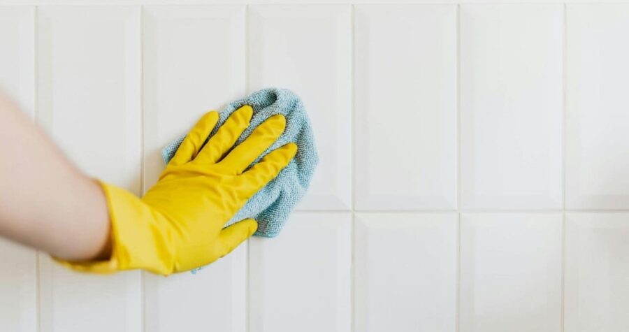 Person wearing a yellow rubber glove wiping white tiled wall with a blue microfiber cloth