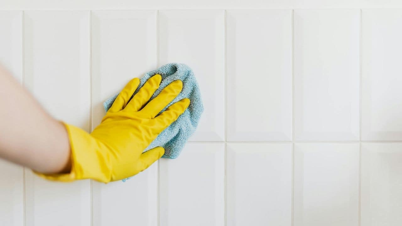 Person wearing a yellow rubber glove wiping white tiled wall with a blue microfiber cloth