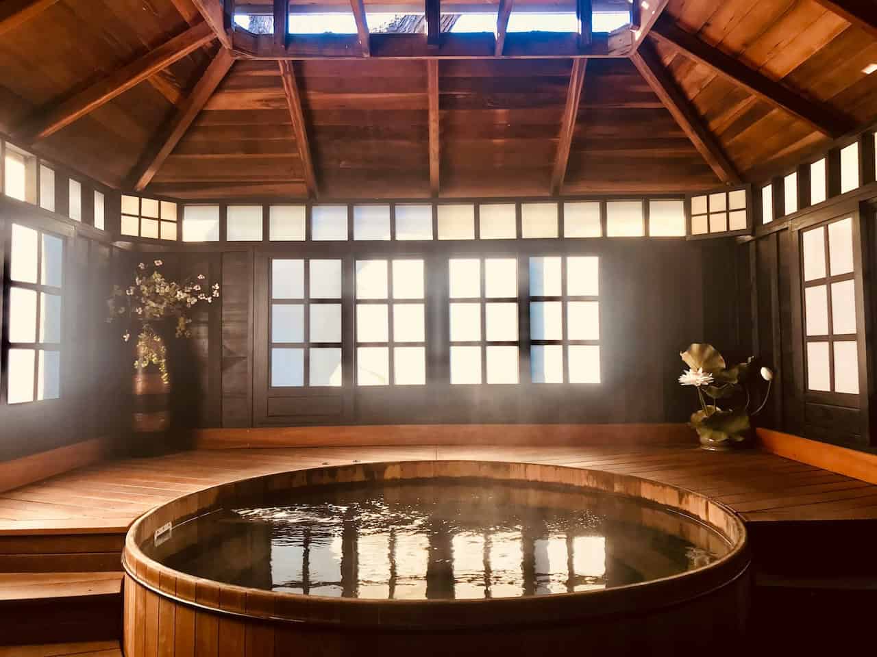 A wooden hot spring bath in a serene interior, natural light streaming through the windows, steam rising from the water, wooden beams and walls, potted plant with a lotus flower beside the bath