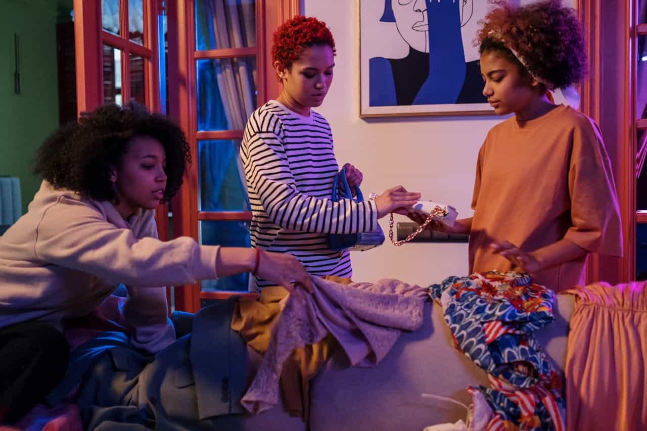 Three young women sorting and exchanging clothes and accessories during a clothes swap party in a cozy room