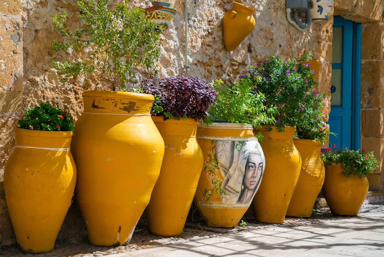 Large yellow ceramic pots and planters arranged against a stone wall, containing various plants including purple flowers and green foliage in a Mediterranean-style garden setting