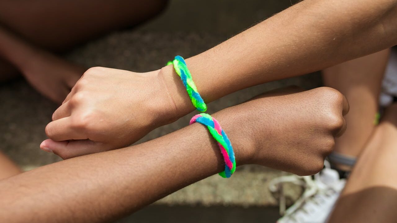 Two hands wearing colorful braided friendship bracelets made from fuzzy pipe cleaners, arms touching in a friendly gesture