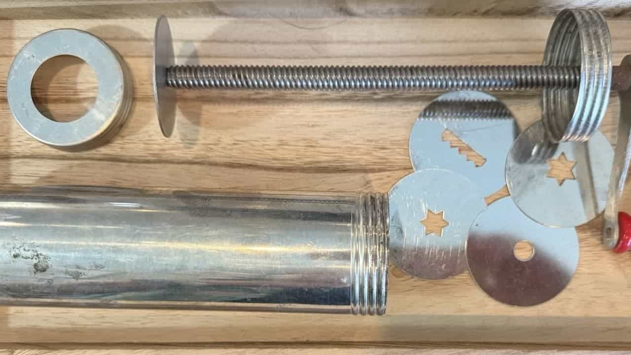 Disassembled metal cookie press on wooden surface, including threaded rod, cylinder barrel, decorative metal discs with star and shape cutouts, and washers