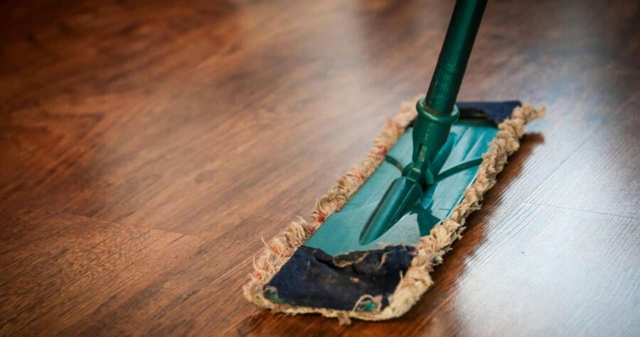 Close-up of a mop cleaning a wooden floor, green handle with a blue and beige mop head, shiny brown wood surface reflecting light