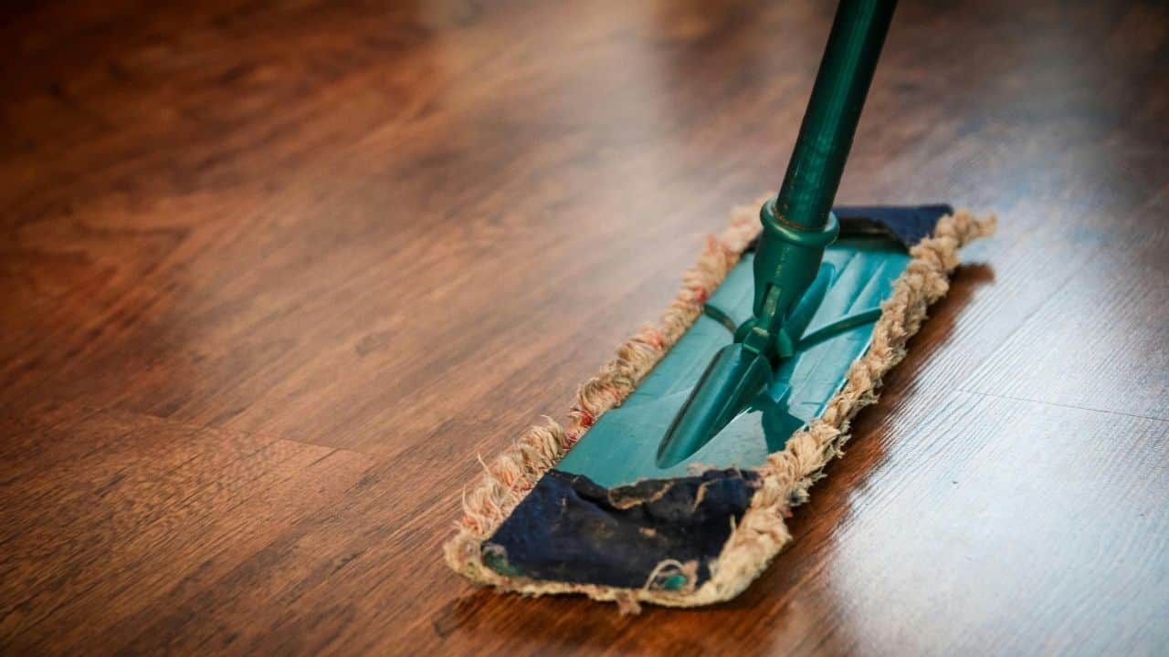 Close-up of a mop cleaning a wooden floor, green handle with a blue and beige mop head, shiny brown wood surface reflecting light
